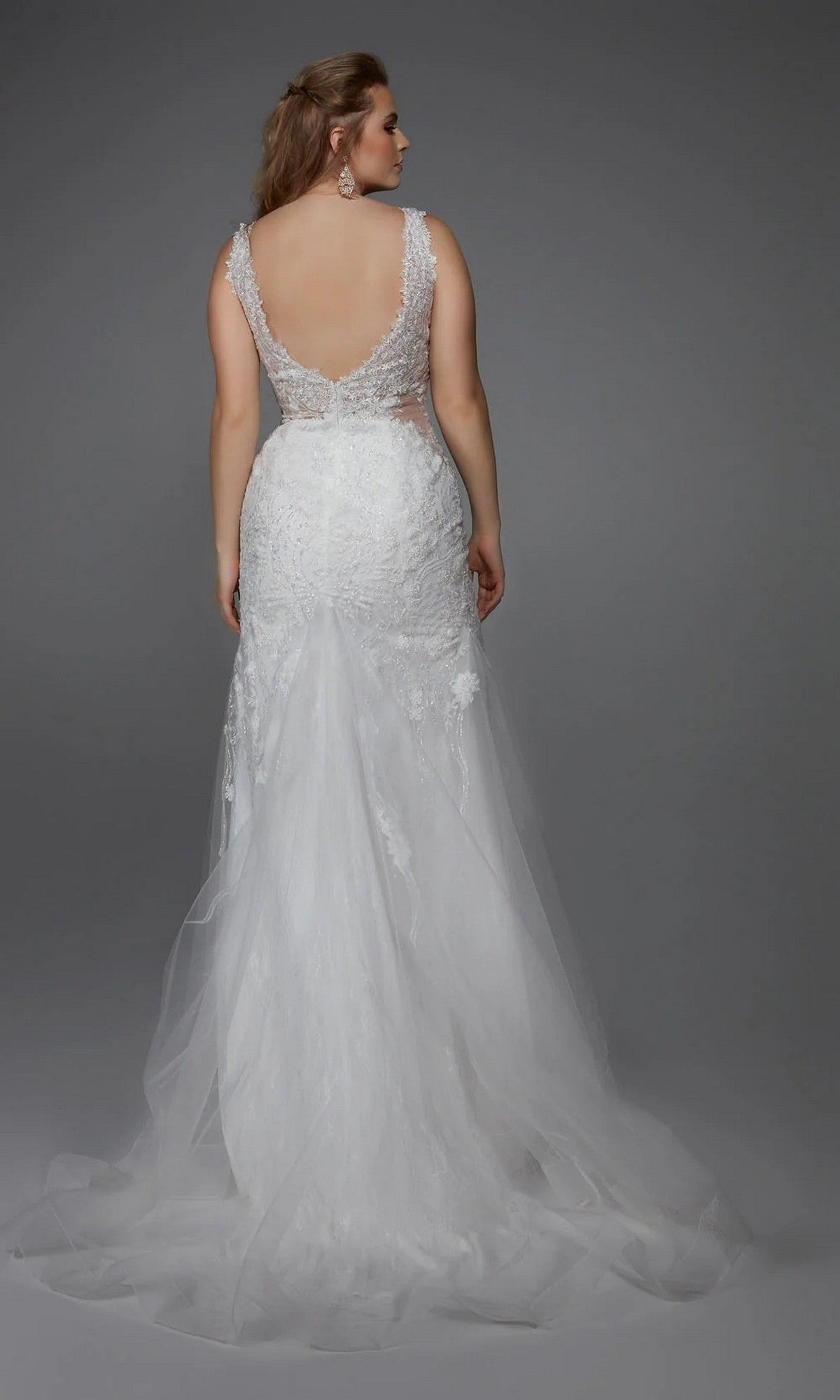 A woman models the Alyce 7017, a long white dress with a lace low back, elegant trumpet skirt, and flowing tulle train, standing against a plain gray background.